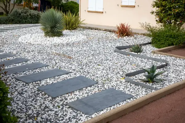 Senderos de piedra gris cruzan jardines con piedras blancas y grises, destacando plantas suculentas y un gran cactus.