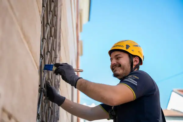 Hombre con casco amarillo sonríe mientras usa un pincel para rebozar paredes y rejas metálicas.