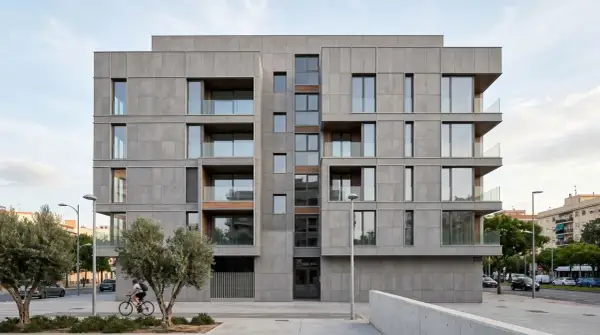 Edificio moderno con una elegante fachada ventilada de paneles grises, balcones acristalados y detalles en madera. Un ciclista pasa frente a árboles y la calle.