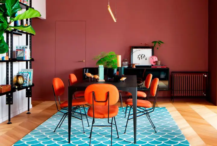 Comedor con paredes color terracota, sillas naranjas vibrantes y una alfombra azul con diseño de escamas. ¡Ideas de colores para pintar un salón audaz!